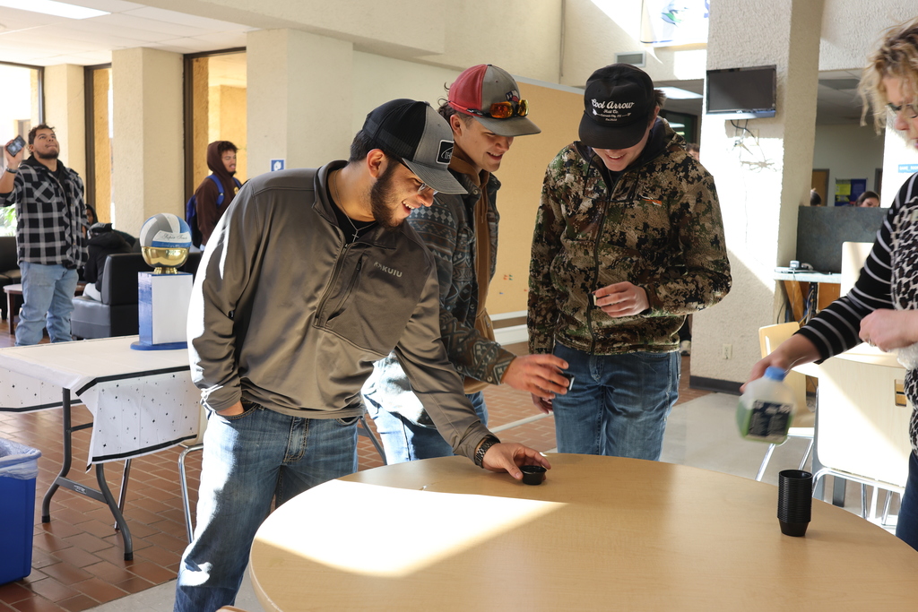 Three men smile standing at the edge of a round table while holding sample cups as a woman holds a jug on the other side.