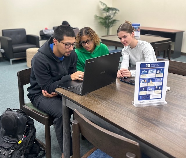A man sitting at at a table looks at a laptop as two women look on next to him.