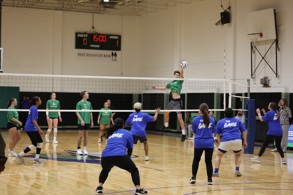 Players in green and blue shirts jump at the net during an active indoor volleyball rally.