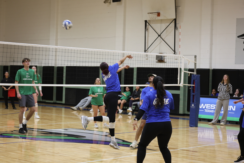 A player in a blue shirt spikes the volleyball while teammates and opponents watch the play unfold.