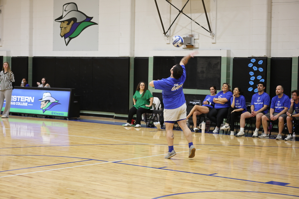 A player in a blue shirt serves a volleyball during an indoor match as teammates watch from the bench.