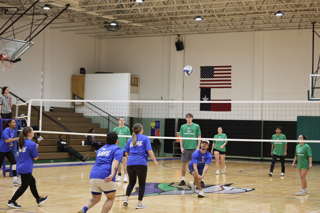 A volleyball flies over the net as students on both teams prepare to receive during a campus game.