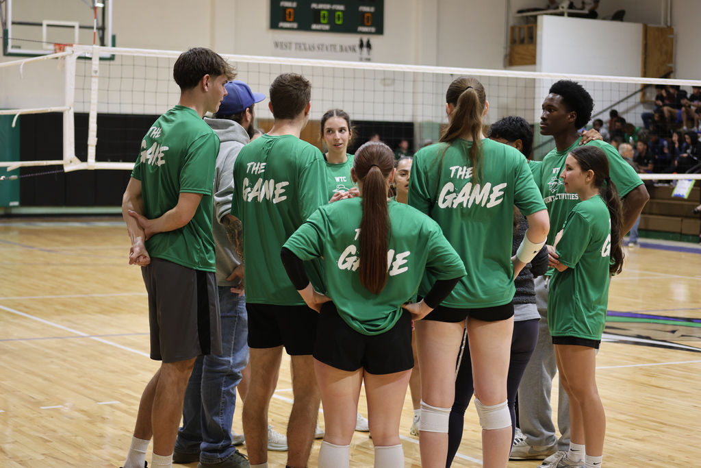 Students wearing green shirts huddle on an indoor volleyball court during a campus recreational game.