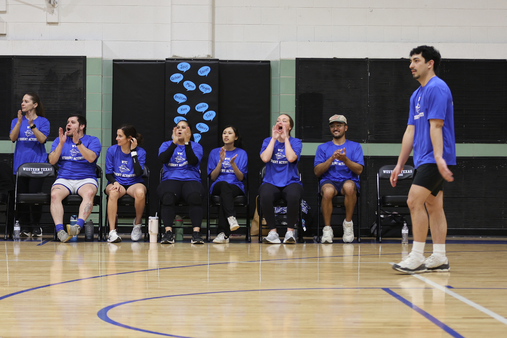 Students in blue shirts cheer and clap from the bench during an indoor volleyball game.