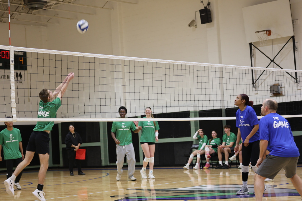 A green-shirted player sends the volleyball over the net as teammates react on the court.