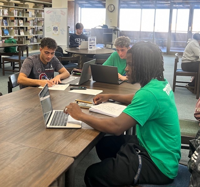 Three men sit at a long table, each with an open laptop and a notebook.