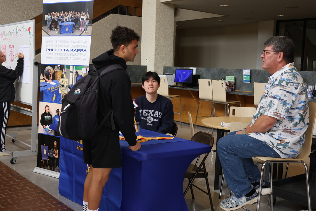 Two men sit at a table while another man leans over to ask a questions.