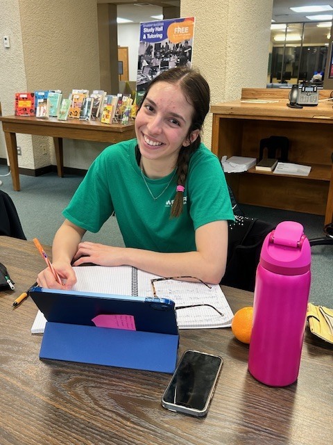A woman smiles while sitting at a table with a pen i her hand and an open laptop in front of her. A pair of glasses sit on a notebook under her elbow with a phone next to the laptop and an orange peeking from behind a tall water bottle.