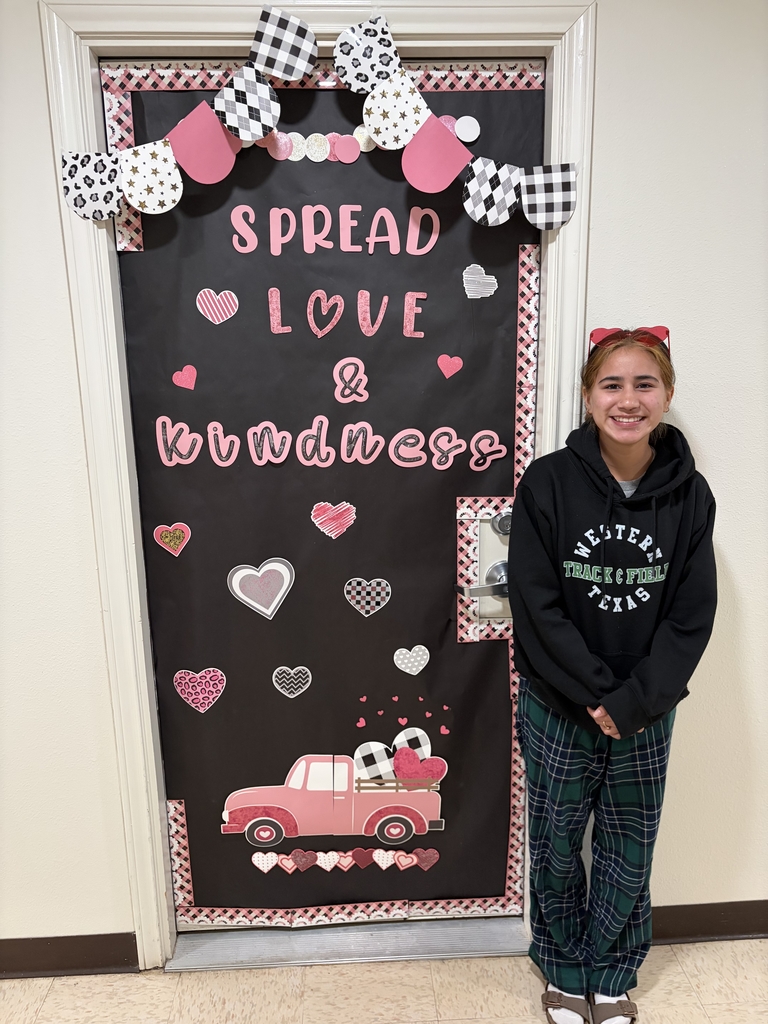 Student smiles beside “Spread Love & Kindness” Valentine’s door decorated with hearts and pink truck.