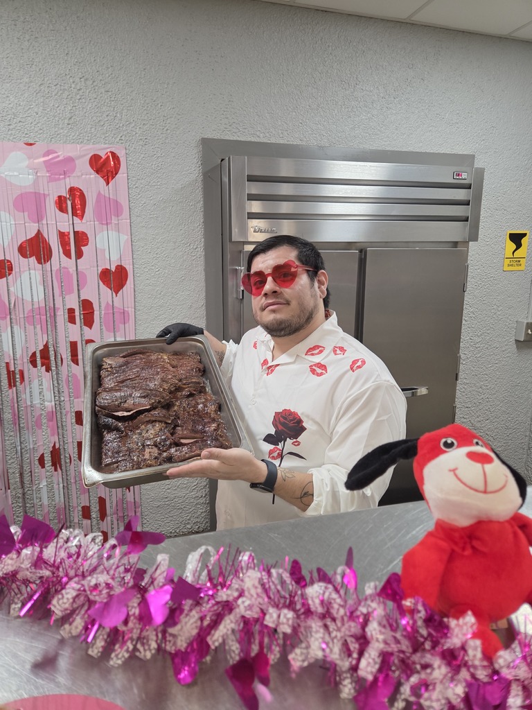 Dining staff member wearing heart glasses holds tray of cooked meat in decorated cafeteria kitchen.