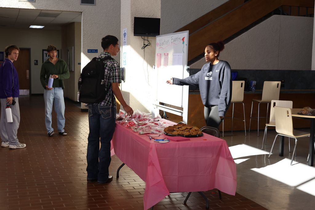 A woman gestures toward Valentine treat table while speaking with a man in campus lobby.