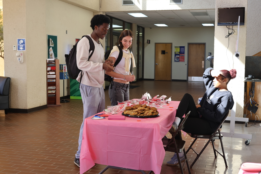 Two students receive Valentine treats from seated volunteer at decorated table.