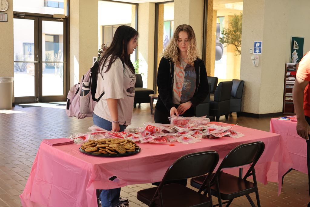 Two students browse Valentine treat bags beside plate of cookies on pink tablecloth.