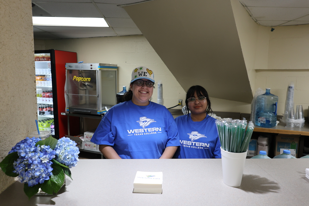 Two PTK members smile behind campus concession counter with popcorn machine and drinks.