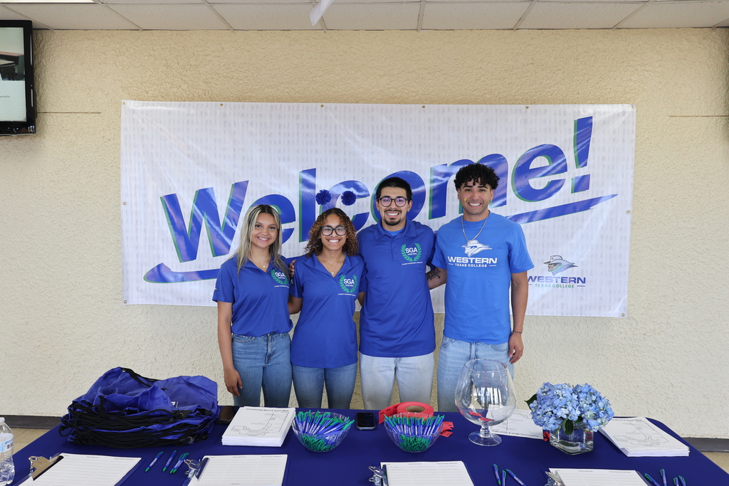 Four Student Government Association members stand at welcome table with pens and bags under welcome banner.