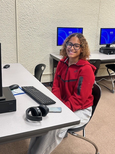 A woman wearing glasses smiles while sitting in front of a computer keyboard with headphones laying next to it.