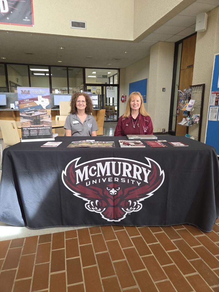 Two women sit at a table covered with a table cloth with the McMurry University logo.