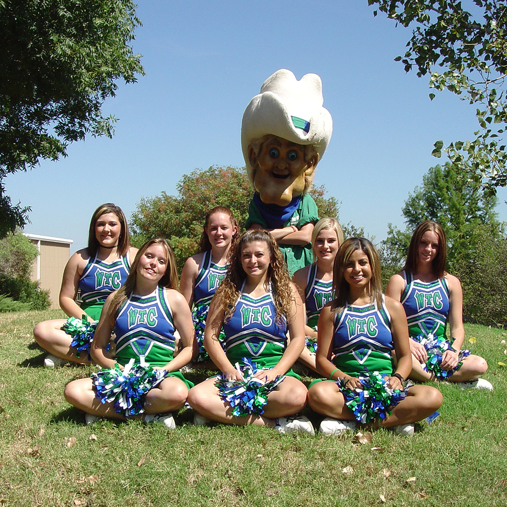 Cheerleaders sit cross legged in the grass with a mascot behind them. 