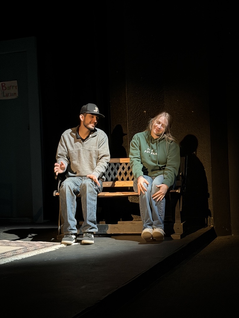 Two actors sit on bench under stage spotlight during Western Texas College production of The Boys Next Door.
