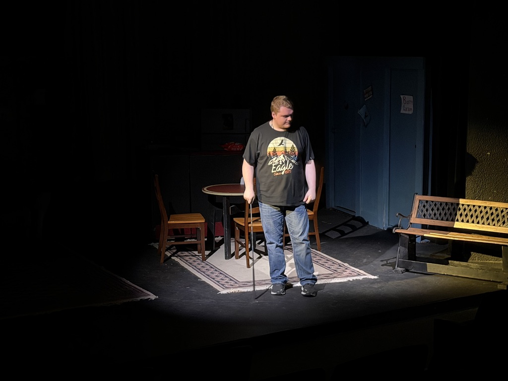 Actor stands alone in spotlight on stage holding a steel rod, kitchen set and bench visible behind him.