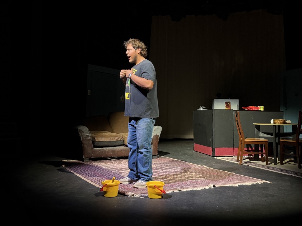 Actor performs on stage beside two yellow cleaning buckets on apartment set during theatrical scene.