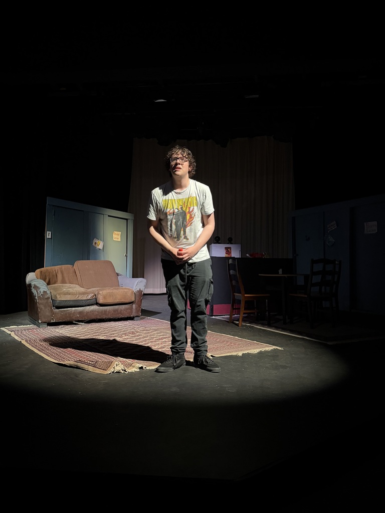 Actor stands center stage in spotlight delivering monologue in living room set with couch and kitchen area behind.