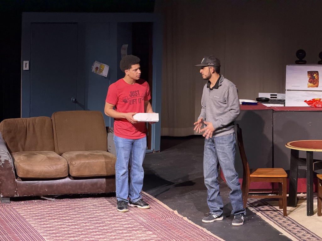 Two actors talk on stage in  a living room/kitchen set, one holding a box while standing near couch and kitchen table.