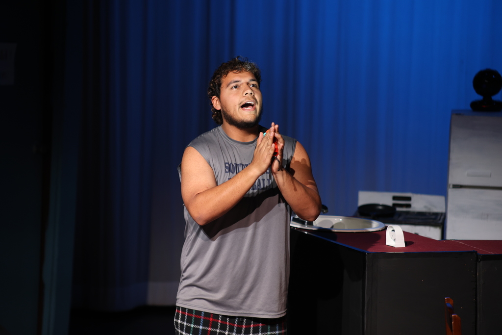 Actor clasps hands in emotional monologue beside kitchen counter under blue stage lighting.