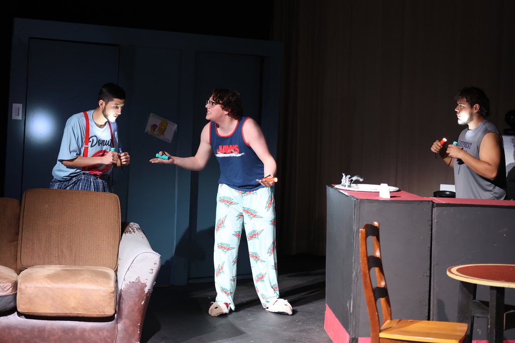 Three actors shine flashlights in apartment set while talking near couch and kitchen counter during stage performance.