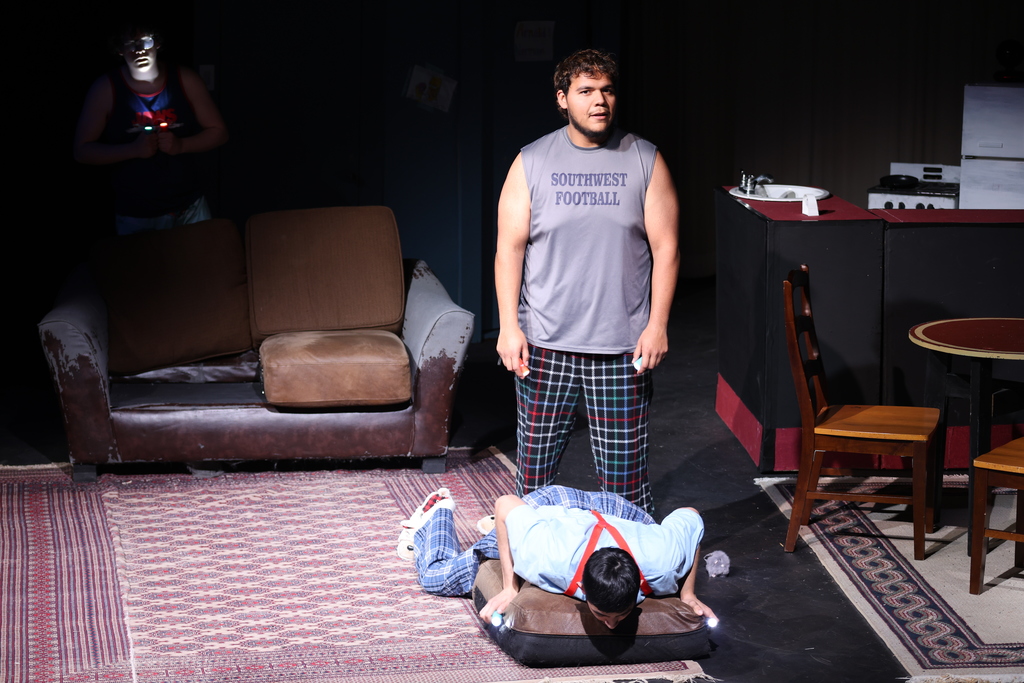 Actor stands while another lies face-down on couch cushion with flashlight during dramatic stage scene.