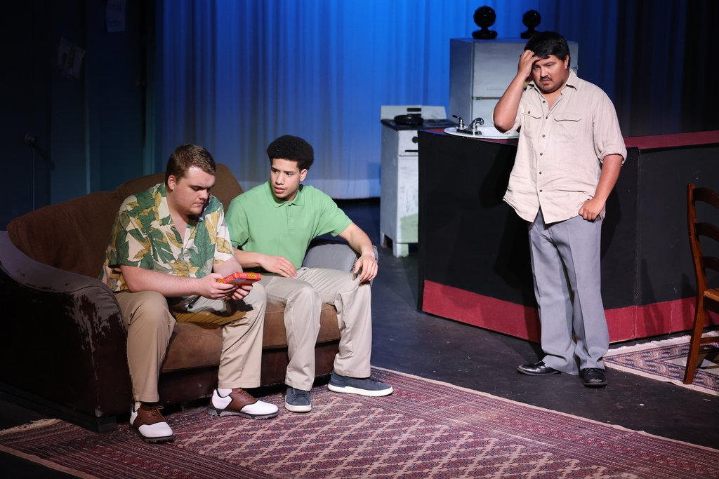 Two actors sit on couch while another stands nearby during tense conversation in apartment set on stage.