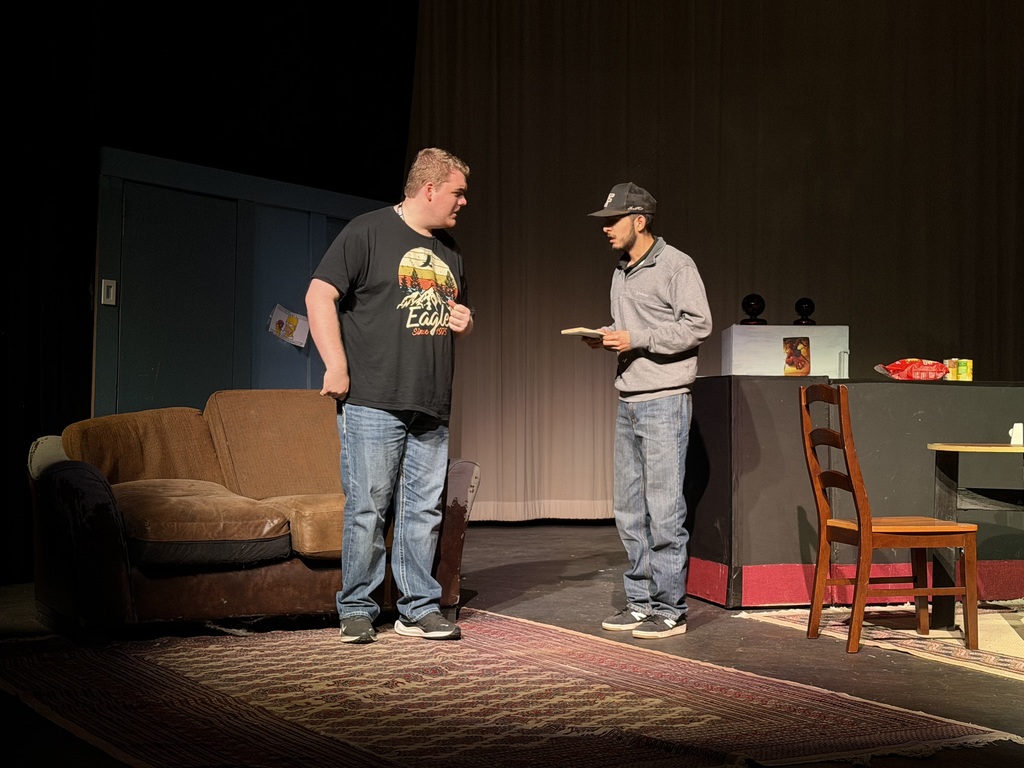 Two actors stand beside couch in living room set discussing something during Western Texas College production of The Boys Next Door.