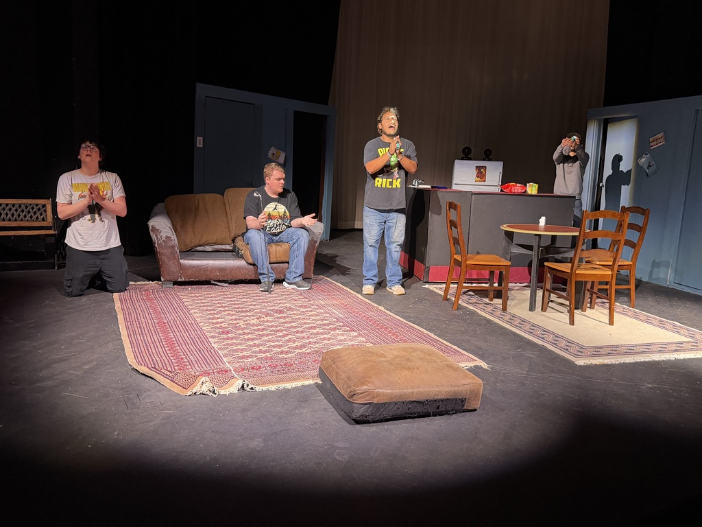 Four actors kneel and pray on stage beside worn couch and kitchen set during Western Texas College production of The Boys Next Door.
