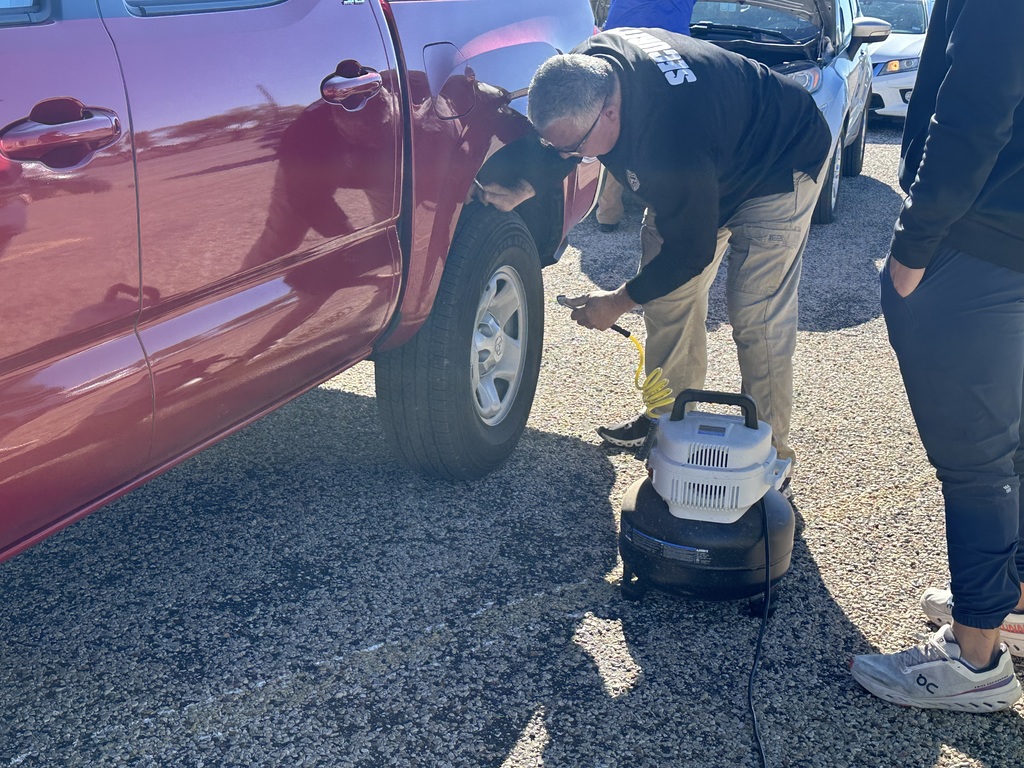 A man looks at a tire pressure gauage standing next to a truck while another man watches.