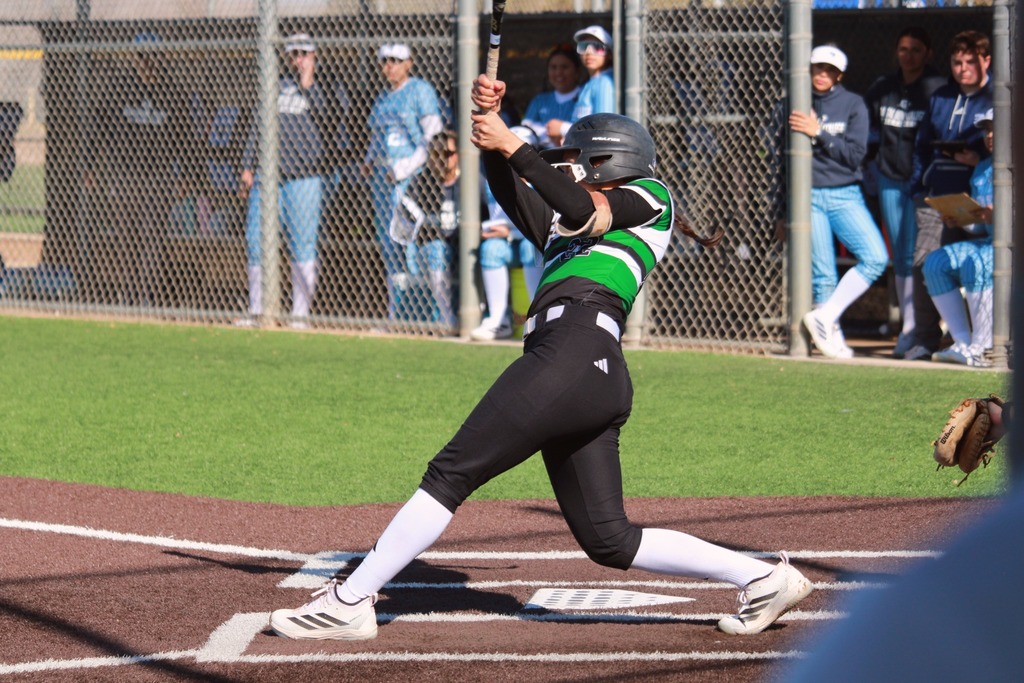 A softball player swings the bat while teammates look on from the dugout.