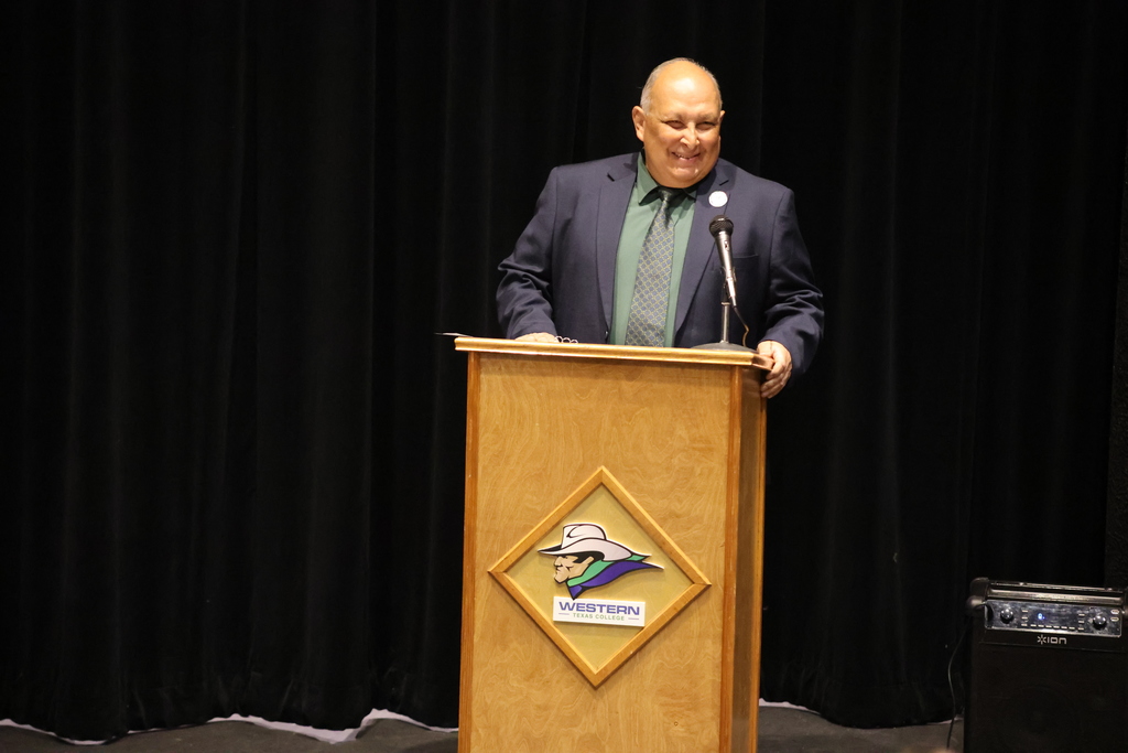 Man speaking at podium with Western Texas College logo during formal event on stage with microphone.