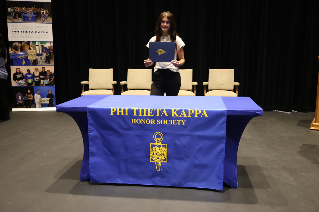 Student holding certificate standing behind Phi Theta Kappa table during induction ceremony on stage.