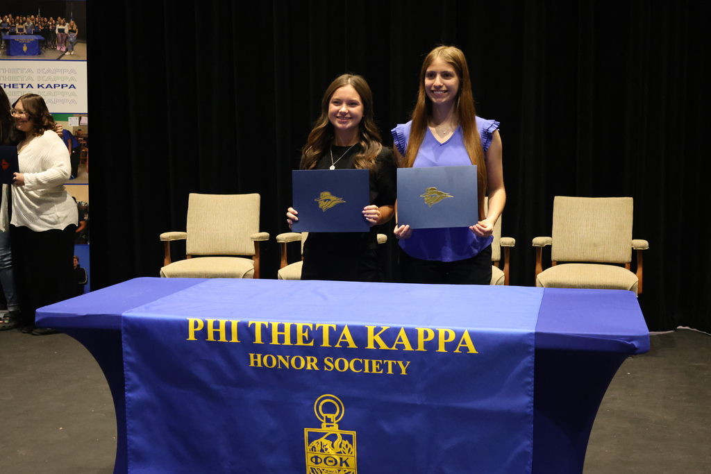 Two students holding certificates and smiling behind Phi Theta Kappa table during induction ceremony on stage.