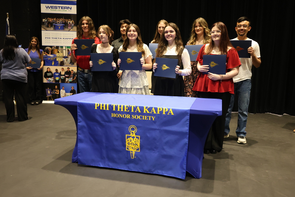 Group of students holding certificates behind Phi Theta Kappa table during induction ceremony with banner display in background.