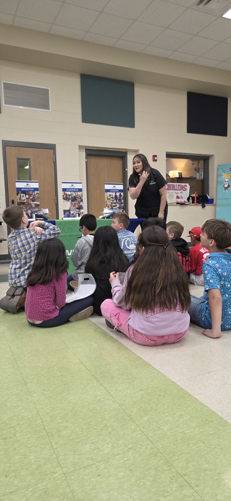 Staff member engages seated elementary students during interactive career day session with WTC information banners in background.