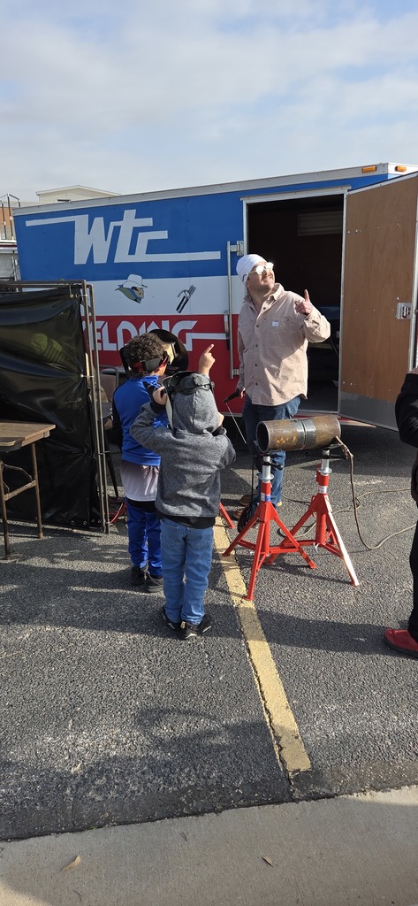 Instructor demonstrates welding setup to children outside WTC trailer while students watch and point at equipment during career day.