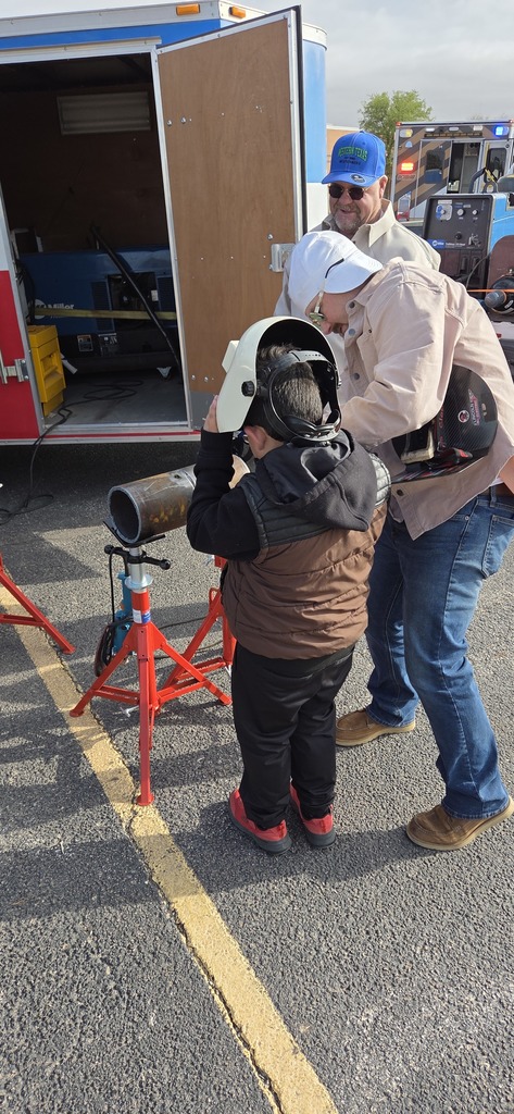 Instructor helps child wear welding helmet and demonstrates pipe welding setup beside WTC trailer during hands on career day activity.