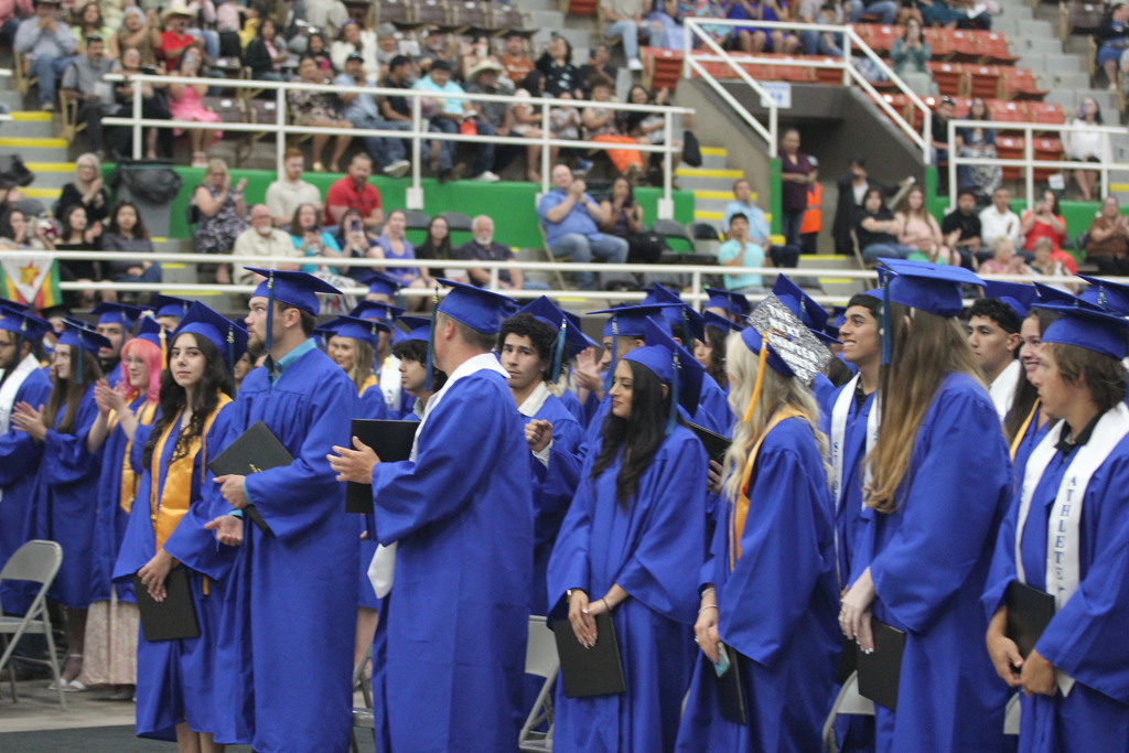 Graduates in blue caps and gowns stand holding diplomas during ceremony with audience seated in arena stands behind them.