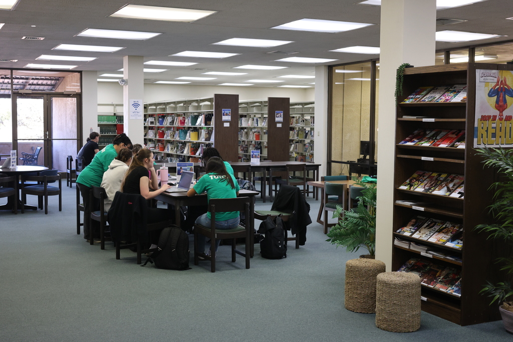 People sit at a table that seats eight with a woman  looking over a person's shoulder behind them. A tall wooden magazine rack stands int he foreground on the right.