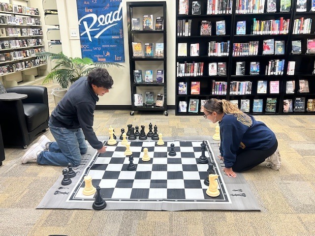 A man and a woman sit across from each other playing chess on an oversized chess board.