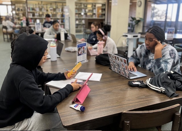 Several women sit at a long table looking at their open laptops and phones.