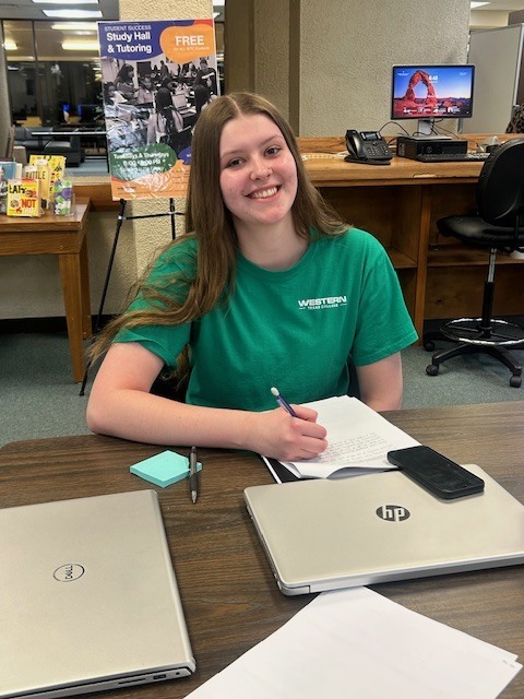 Student in green shirt writes notes at table with laptops during study hall.