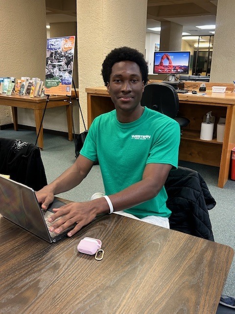 Student in green shirt uses laptop at library table with phone and earbuds nearby.