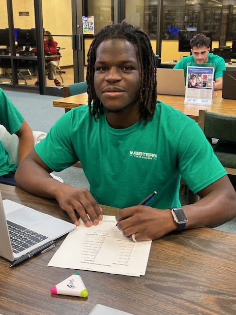 Student in green shirt writes on worksheet beside laptop in study hall.