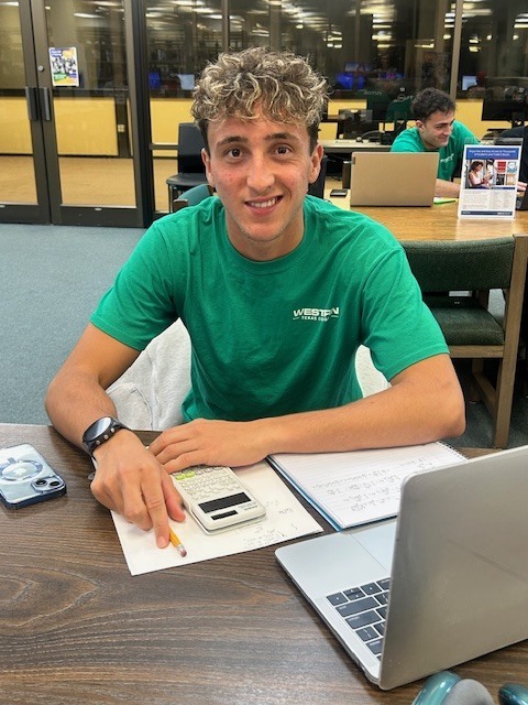 Student in green shirt studies with calculator and notebook at library table.